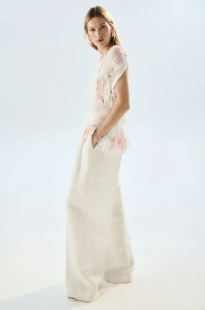Model Liz Kennedy wearing wide-leg cream trousers and a pleated floral sleeveless blouse against a clean white backdrop.