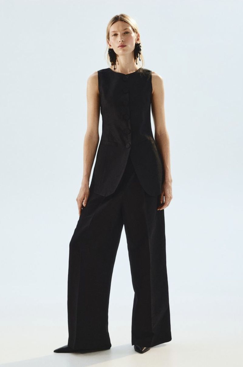 Model Liz Kennedy wearing a black sleeveless tailored longline vest and wide-leg trousers against a white studio background.