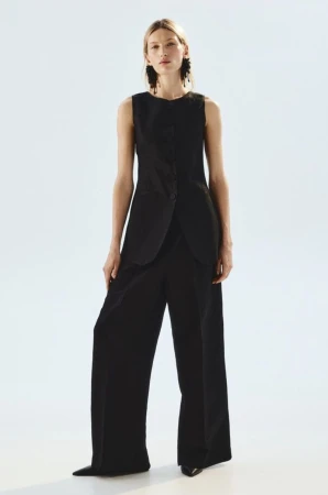 Model Liz Kennedy wearing a black sleeveless tailored longline vest and wide-leg trousers against a white studio background.