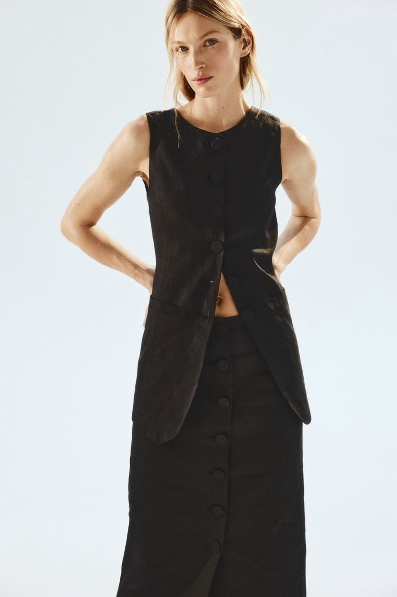 A fashionable woman poses against a white background in a sleeveless black linen top and matching long skirt with buttons down the front.
