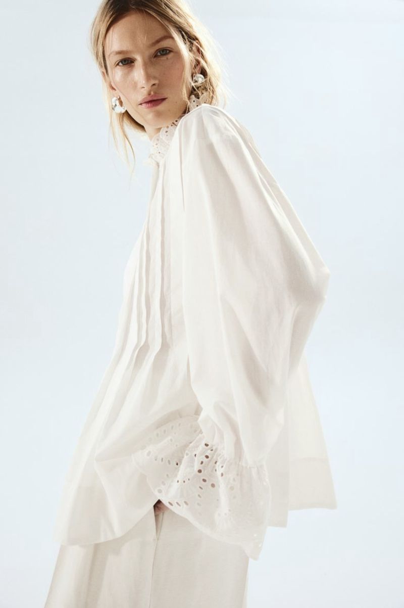 A woman in a white blouse and pants poses in a studio setting with soft lighting and a light blue background, showcasing fashion apparel.