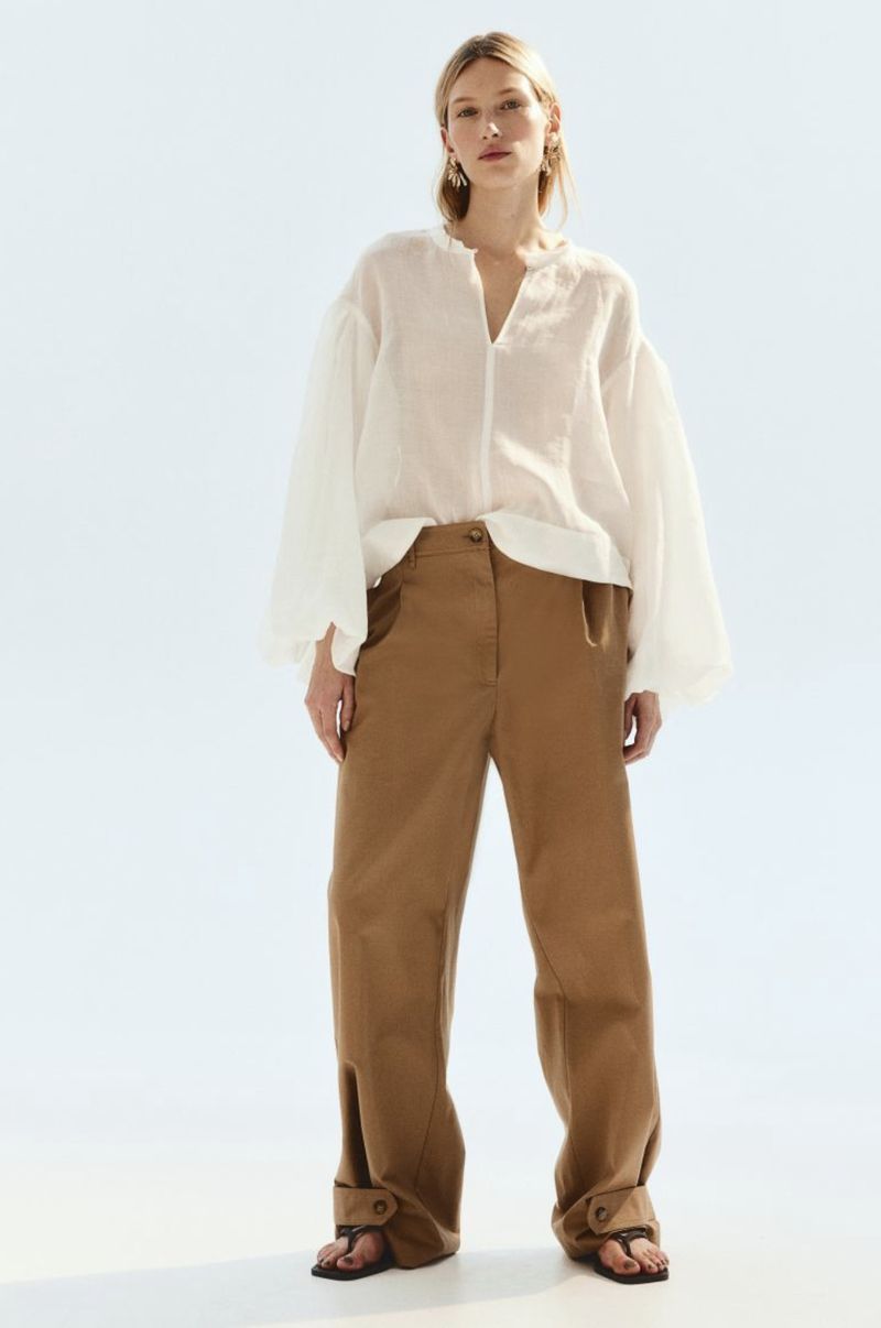 A fair-skinned young adult woman poses in a studio wearing a sheer white blouse with large sleeves and tan, wide-legged trousers with sandals.