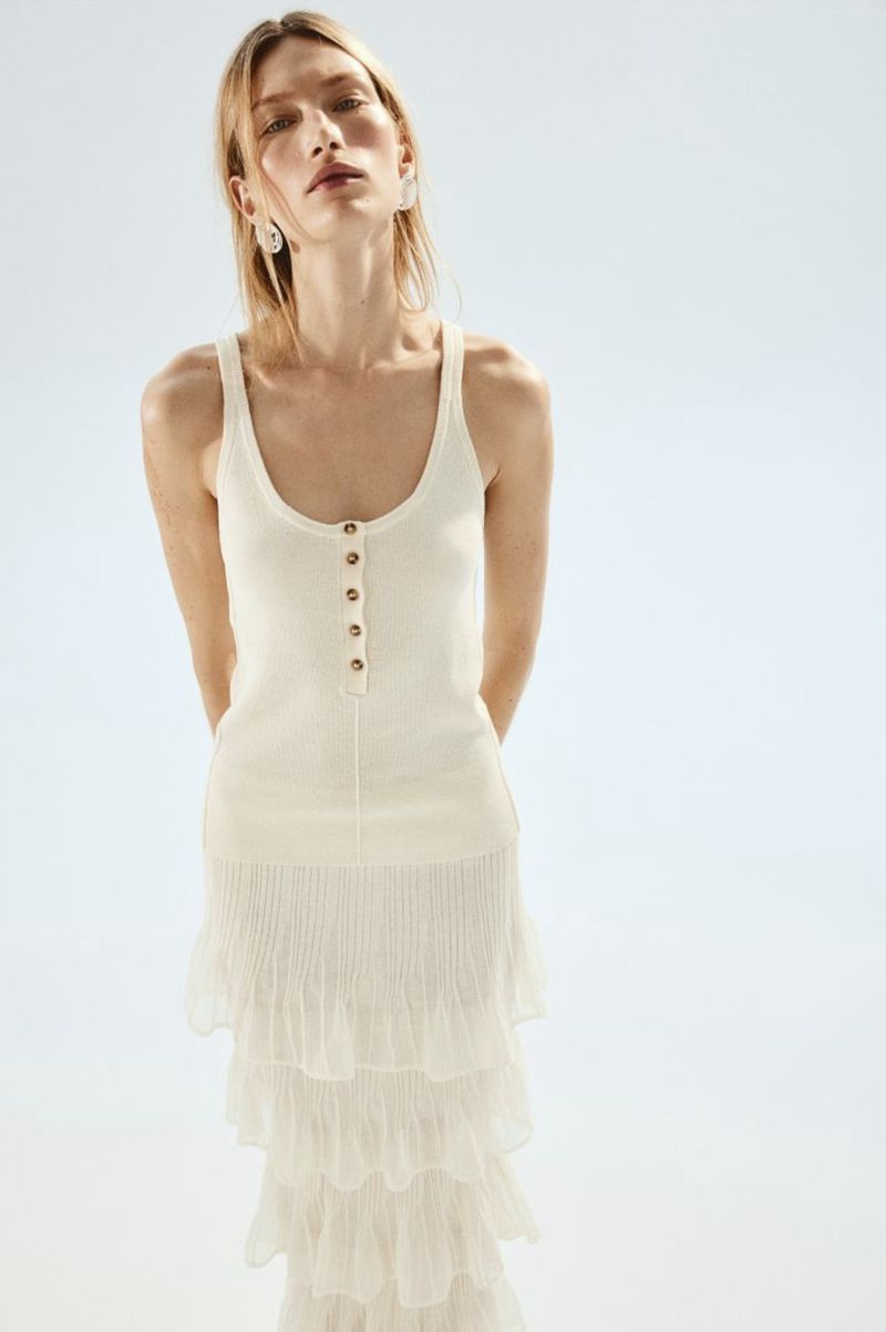 A blonde woman poses in a studio wearing an off-white knit top with buttons and a tiered ruffled skirt against a light background.