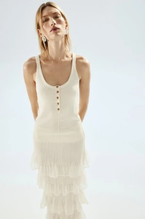 A blonde woman poses in a studio wearing an off-white knit top with buttons and a tiered ruffled skirt against a light background.