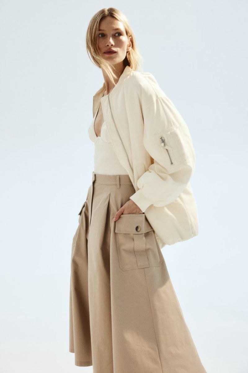Adult female fashion model in a studio wearing a white bomber jacket and tan cargo skirt, posing in front of a bright background.