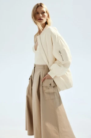 Adult female fashion model in a studio wearing a white bomber jacket and tan cargo skirt, posing in front of a bright background.