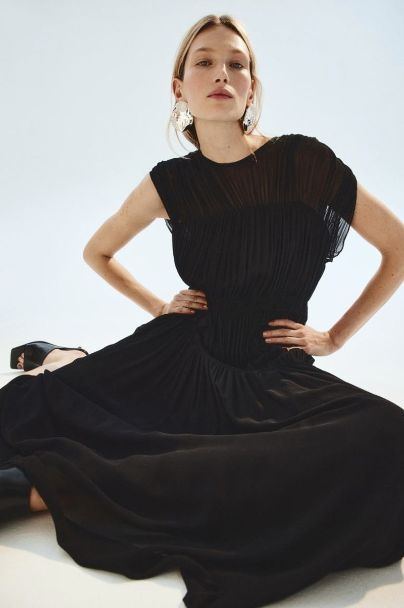 Woman in elegant black maxi dress poses with hands on her waist in a studio with a clean white background.