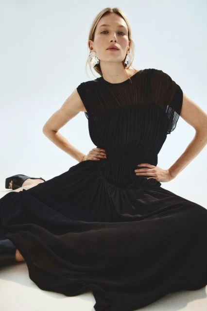 Woman in elegant black maxi dress poses with hands on her waist in a studio with a clean white background.
