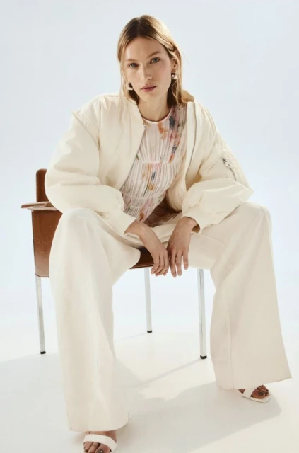 A woman sits in a chair wearing wide-leg pants, sandals, and a bomber jacket in a fashion studio shoot with a white backdrop.