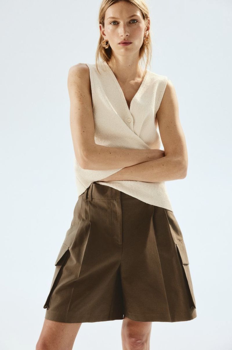 Woman in sleeveless sweater and olive cargo shorts poses in a studio. The fashion shot has bright lighting and a medium shot.