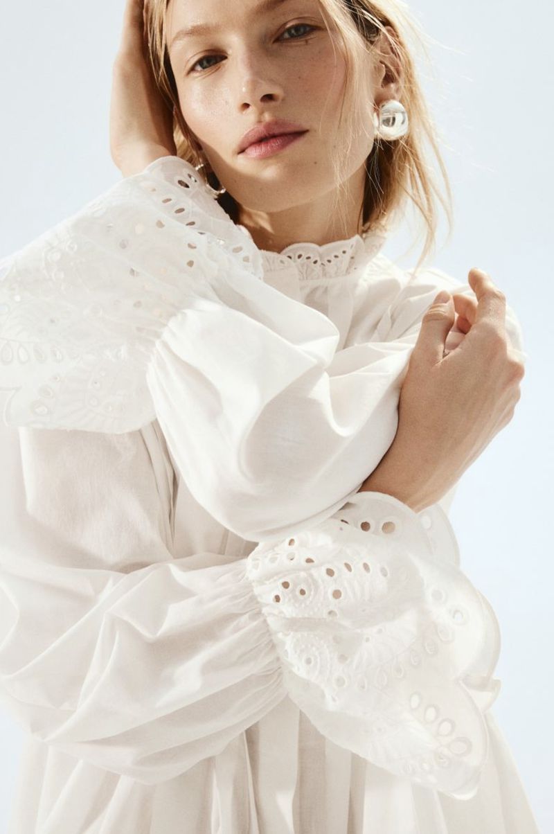 Blond woman poses in a white eyelet long-sleeved blouse against a bright background, showcasing fashion and beauty elements.
