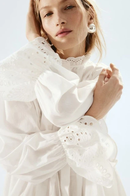 Blond woman poses in a white eyelet long-sleeved blouse against a bright background, showcasing fashion and beauty elements.