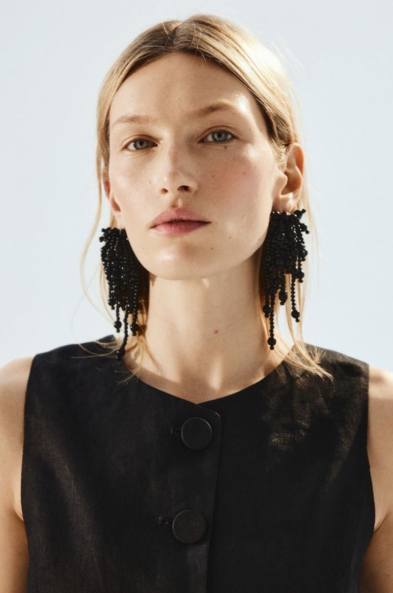 Close-up of a blonde woman with large black beaded earrings wearing a black top against a white background in a studio.