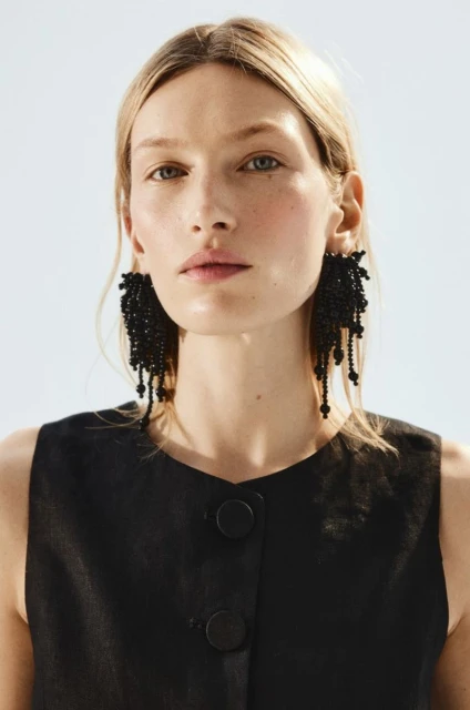 Close-up of a blonde woman with large black beaded earrings wearing a black top against a white background in a studio.
