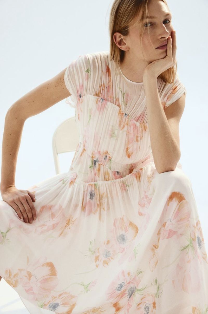 A fair-skinned woman with blonde hair poses in a light floral dress, seated with a thoughtful expression in a bright studio setting.
