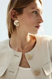 Model wearing a light-coloured jacket with large metallic buttons and round earrings against a clear sky