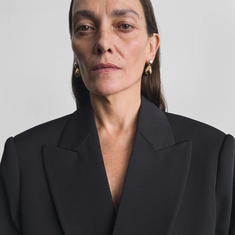 Close-up portrait of a woman with dark hair wearing a black blazer and gold earrings, set against a plain white background.