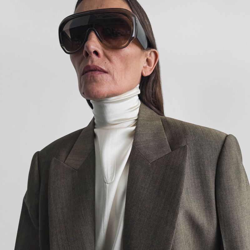 An adult female with dark hair wears large sunglasses, a white turtleneck shirt, and a dark blazer against a neutral background.