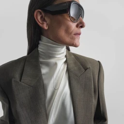 Laura Ponte in large dark shield sunglasses, an ivory silk turtleneck top, and a tailored wool blazer against a seamless studio backdrop.