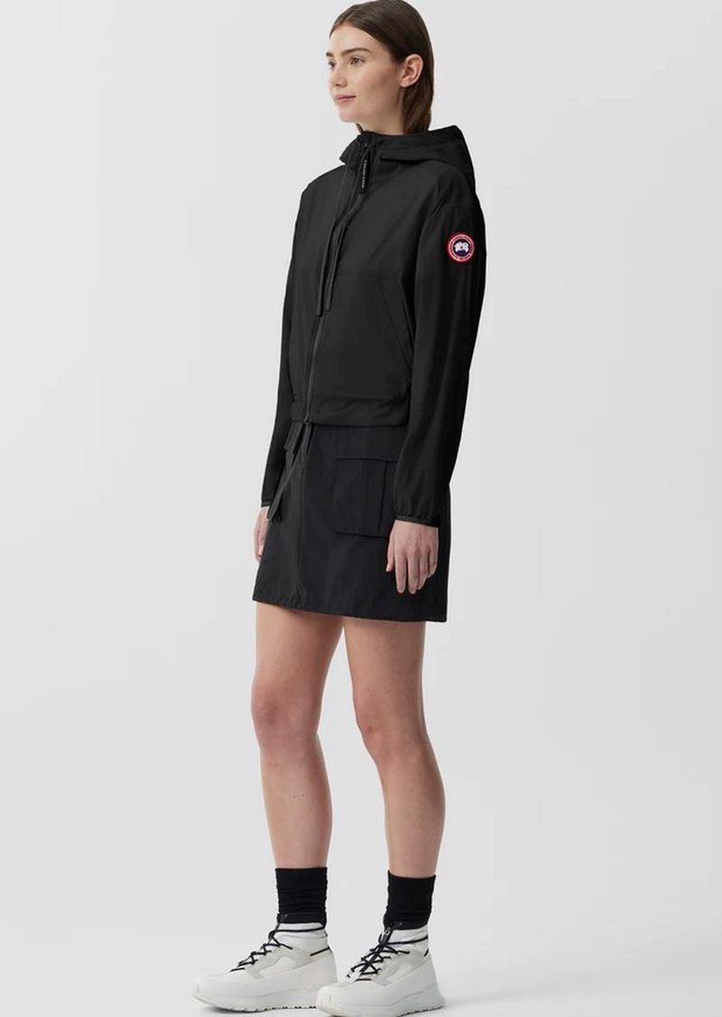 A full-length shot of an adult woman wearing a black hooded jacket and a black skirt, paired with white sneakers, set against a white background.