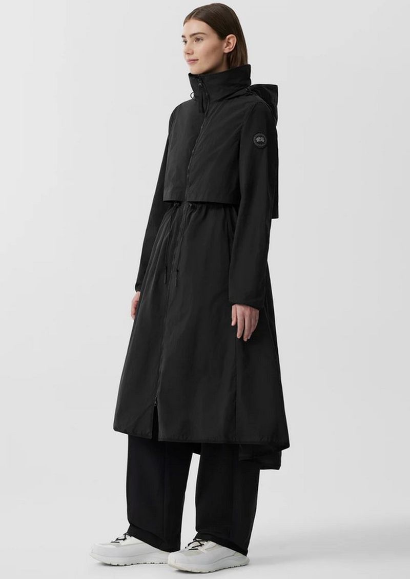 A woman models a stylish, long black raincoat, paired with wide black pants and white sneakers, against a clean, bright background.