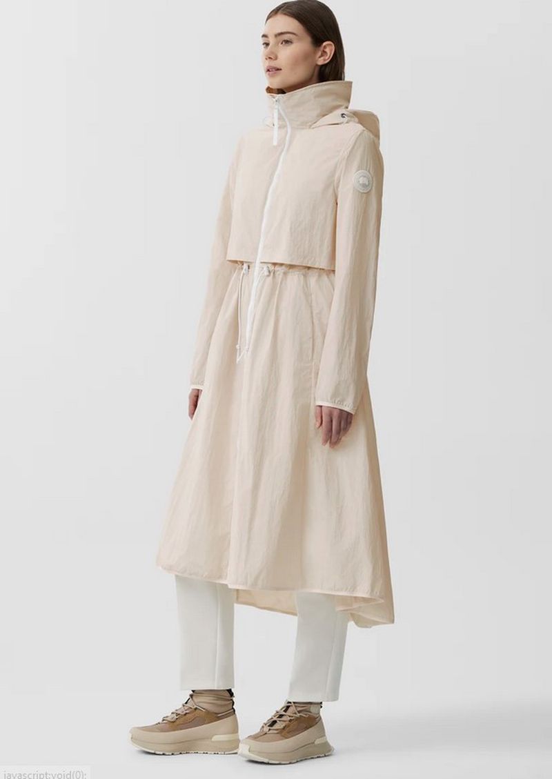 Fashion shot of a woman in a long, light coat with a hood, white pants, and light-brown shoes against a plain white backdrop.