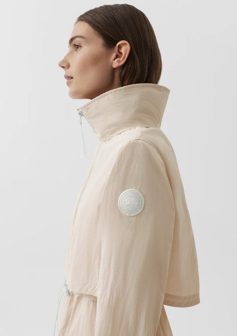 Profile view of an adult female wearing a light pink jacket with a high collar, against a solid off-white backdrop in a studio setting.
