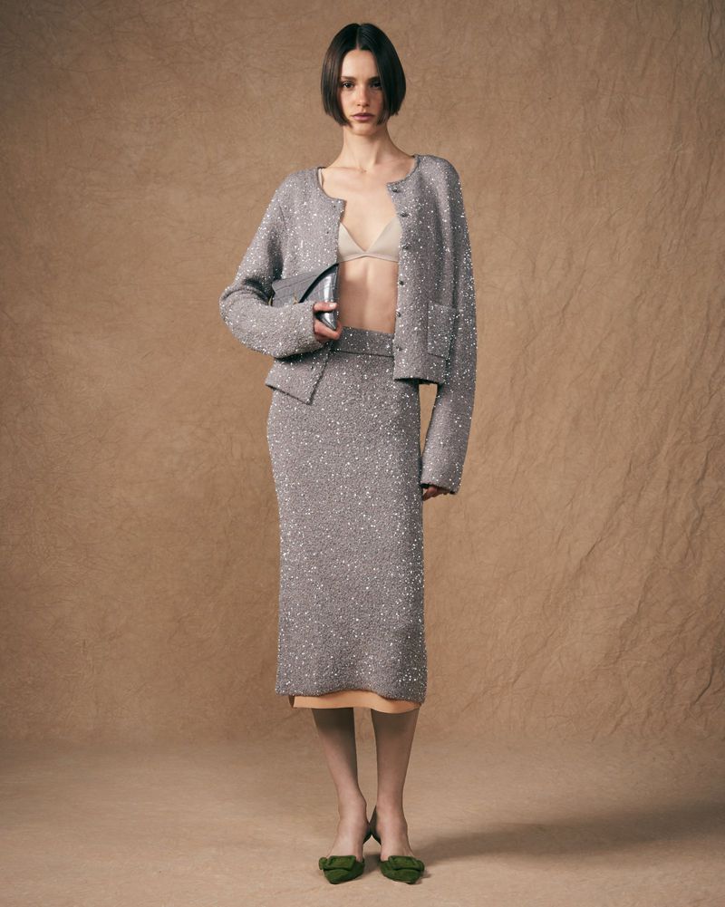 A woman in a gray, sparkly knit cardigan and pencil skirt poses in a studio with a neutral background, holding a metallic purse.