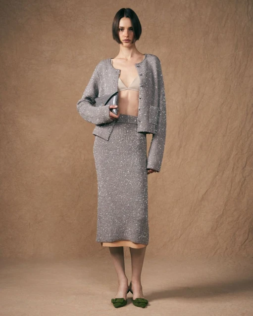 A woman in a gray, sparkly knit cardigan and pencil skirt poses in a studio with a neutral background, holding a metallic purse.