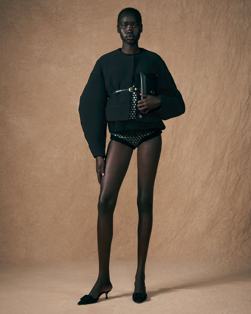 Full-length studio shot of an African American woman in black jacket, patterned bottoms, and heels posing in front of beige background.