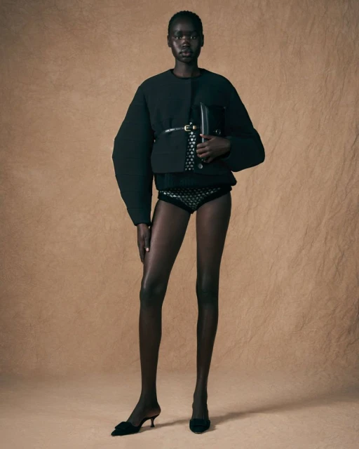 Full-length studio shot of an African American woman in black jacket, patterned bottoms, and heels posing in front of beige background.