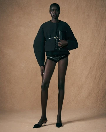 Full-length studio shot of an African American woman in black jacket, patterned bottoms, and heels posing in front of beige background.