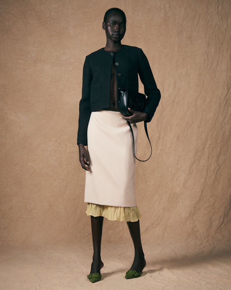 Full-length shot of a Black woman modeling a black jacket, cream skirt, and black bag in a studio setting. Fashion editorial.