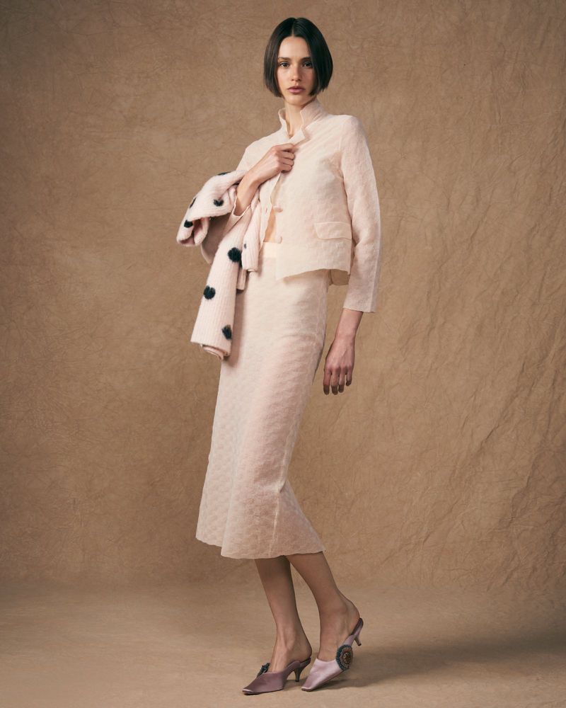 Elegant woman in a light pink patterned skirt suit with retro heeled mules poses in a fashion studio against a textured background.