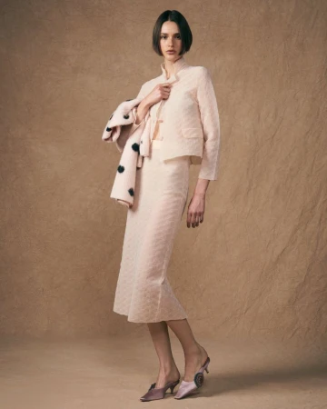 Elegant woman in a light pink patterned skirt suit with retro heeled mules poses in a fashion studio against a textured background.