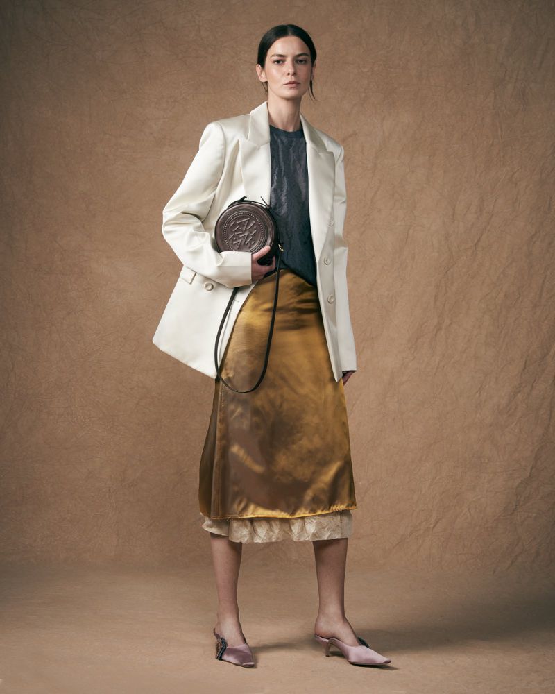 Fashion portrait of woman in oversized white blazer and gold skirt holding round brown handbag against a neutral brown backdrop.