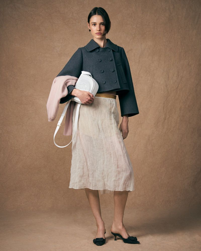 A woman in a short grey jacket, beige skirt, and black heeled mules stands in front of a mottled brown backdrop holding a white purse.