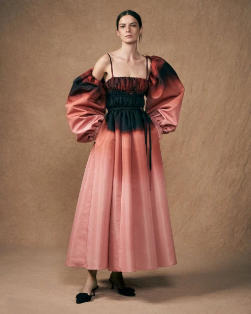 Elegant woman in a light pink couture gown with black accents, posing against a textured beige backdrop in a fashion forward portrait.