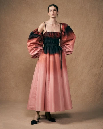 Elegant woman in a light pink couture gown with black accents, posing against a textured beige backdrop in a fashion forward portrait.