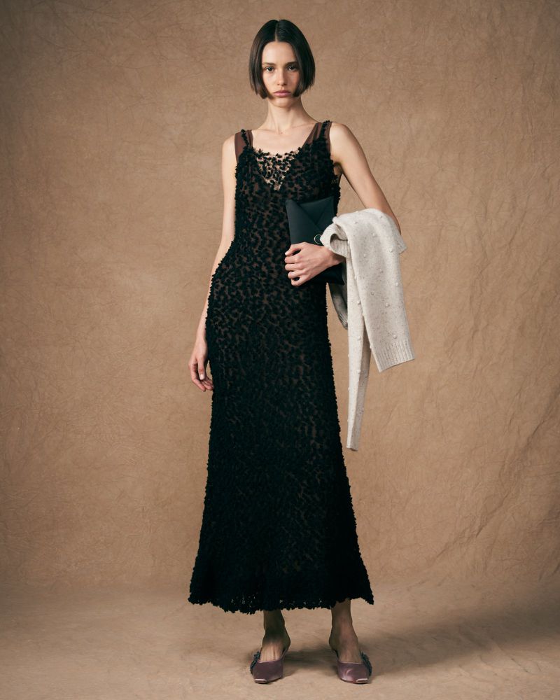 An adult woman stands in a studio setting, wearing a black evening dress and holding a clutch and shawl, presented in a fashion-editorial style.