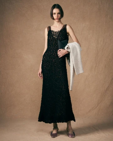 An adult woman stands in a studio setting, wearing a black evening dress and holding a clutch and shawl, presented in a fashion-editorial style.