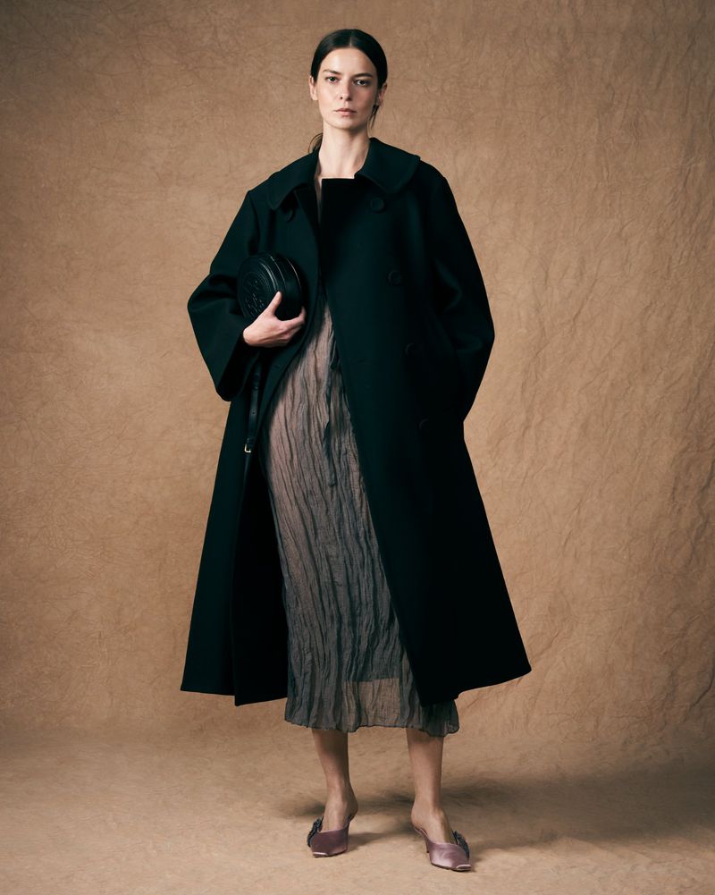 A woman stands in a studio wearing a long black coat, a sheer dress, and low satin heels, holding a round black purse.