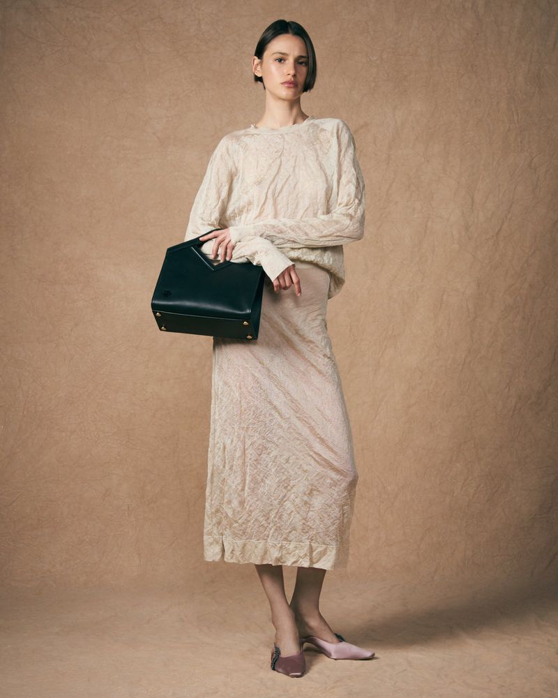 Adult female model poses in a long skirt and sweater, holding a black purse in a studio with a neutral backdrop for a fashion shot.