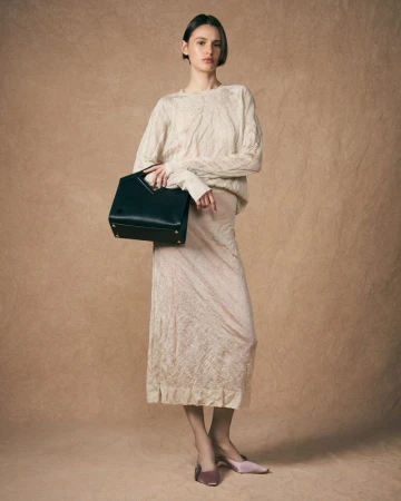 Adult female model poses in a long skirt and sweater, holding a black purse in a studio with a neutral backdrop for a fashion shot.