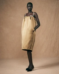 Model standing against a textured brown backdrop wearing a voluminous gold satin dress and black heels.