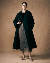 Model in dark coat and textured midi dress holding a black handbag against a textured brown backdrop.
