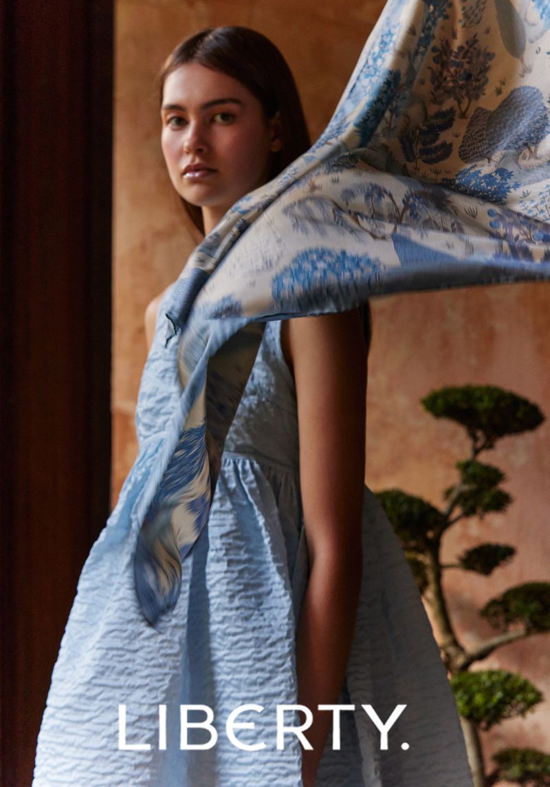 A young adult woman poses indoors wearing a blue dress and a printed shawl or scarf, with soft lighting and a decorative houseplant.