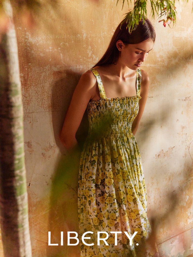 Young woman in a yellow floral print dress stands near a textured wall with shadows and a glimpse of green foliage overhead.