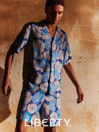 A young adult male poses indoors wearing a blue floral shirt and matching shorts, with a textured background and the LIBERTY logo.
