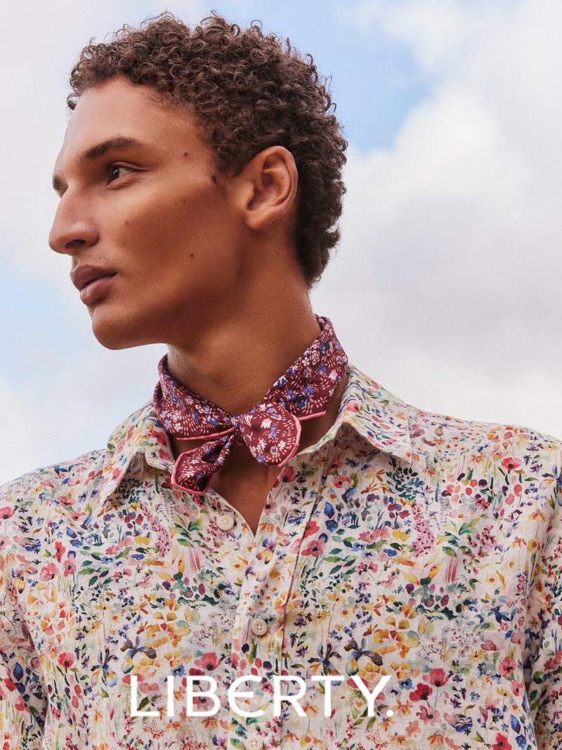 A curly-haired man in a floral shirt with a floral neck scarf poses against a blue sky. Fashion editorial photography.
